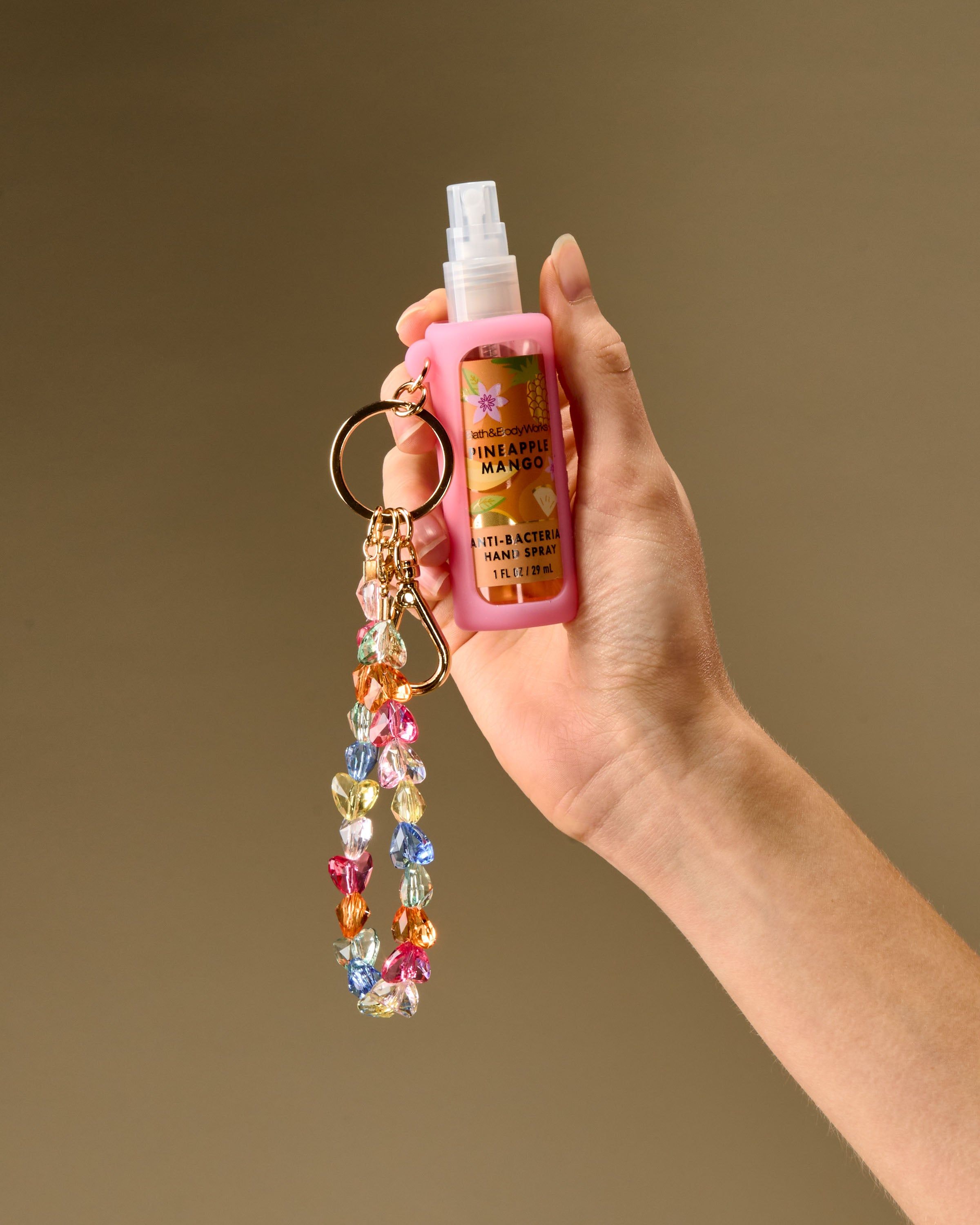 Różowy holder na spray z kolorową bransoletką‑brelokiem i złotym karabińczykiem, idealny do przypięcia w podróży.